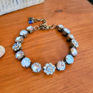 May include: A vintage-style bracelet with iridescent, light blue, and clear crystal-like stones. The bracelet has a bronze-colored chain and settings. The design includes a small blue charm and a floral-shaped stone. The bracelet is on a wooden surface.