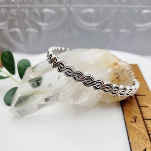 May include: A silver bangle bracelet with a braided design. The bracelet is on a white crystal and a wooden ruler.