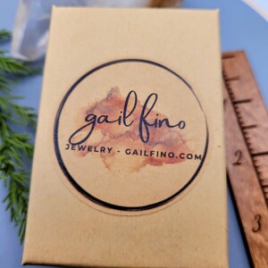 以下が含まれることがあります： 黒い文字で「gail fino JEWELRY - GAILFINO.COM」と書かれた丸いステッカーが付いた、小さな茶色の段ボール箱です。
