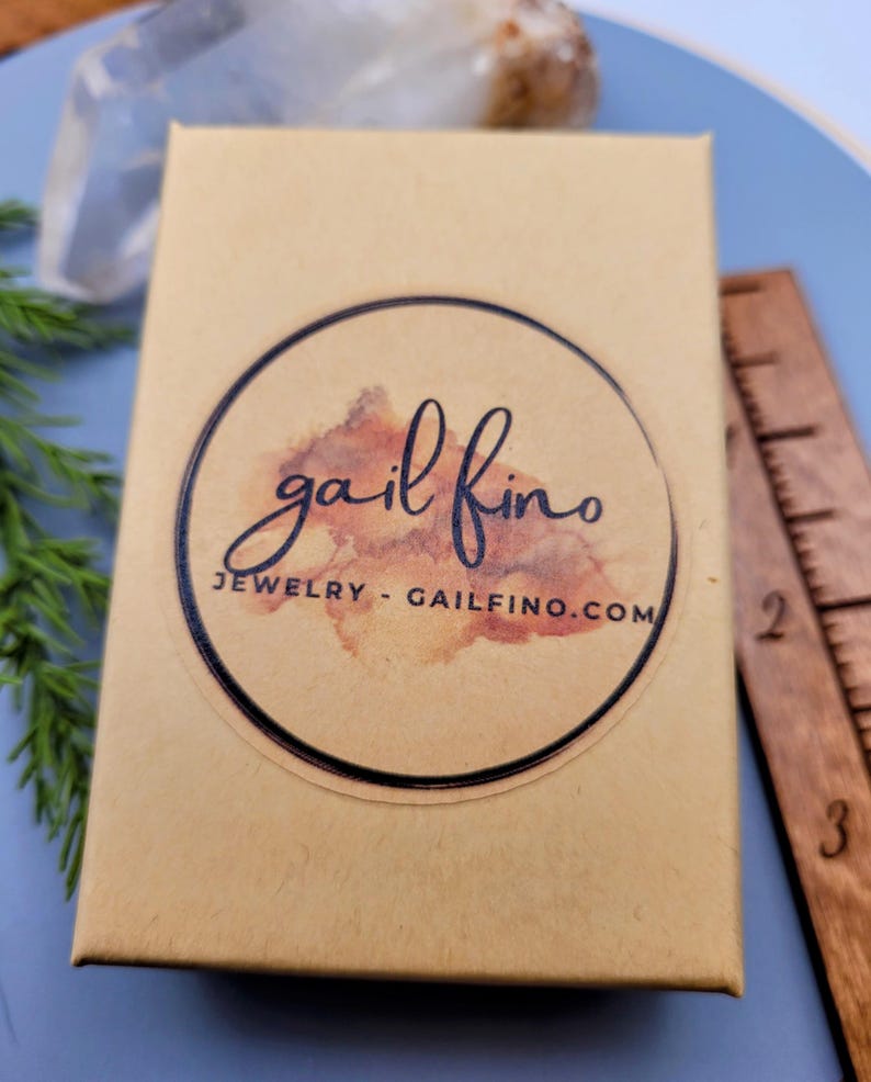 以下が含まれることがあります： 黒い文字で「gail fino JEWELRY - GAILFINO.COM」と書かれた丸いステッカーが付いた、小さな茶色の段ボール箱です。