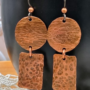 May include: A pair of copper earrings with a hammered texture. Each earring features a circular top and a rectangular bottom, connected by a small copper loop. The earrings hang from a silver-colored hook with a small copper bead.