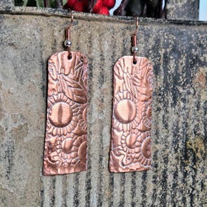 May include: A pair of copper-colored earrings with a rectangular shape. The earrings feature an embossed floral and circular pattern. The earrings hang from copper-colored ear wires with a small silver bead.