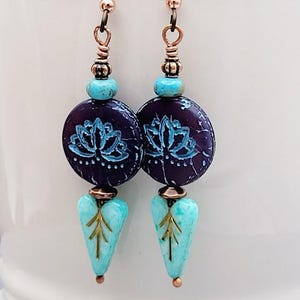 May include: A pair of handcrafted earrings featuring dark purple, turquoise, and copper-colored beads. The earrings have a lotus flower design on the purple beads and leaf-shaped turquoise pendants. The ear wires and connecting beads are copper-colored.
