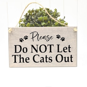 May include: A rectangular wooden sign with the text "Please Do NOT Let The Cats Out" in black. The sign has paw prints and a small green plant on top, with a twine hanger.