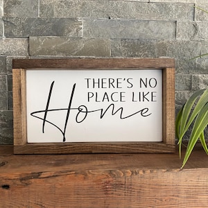 May include: A wooden sign with a white background and black text that reads "There's no place like home". The sign is framed in a dark brown wood frame.