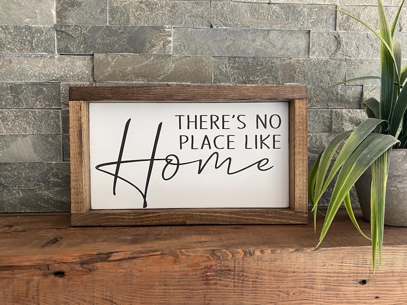 May include: A wooden sign with a white background and black text that reads "There's no place like home". The sign is framed in a dark brown wood frame.