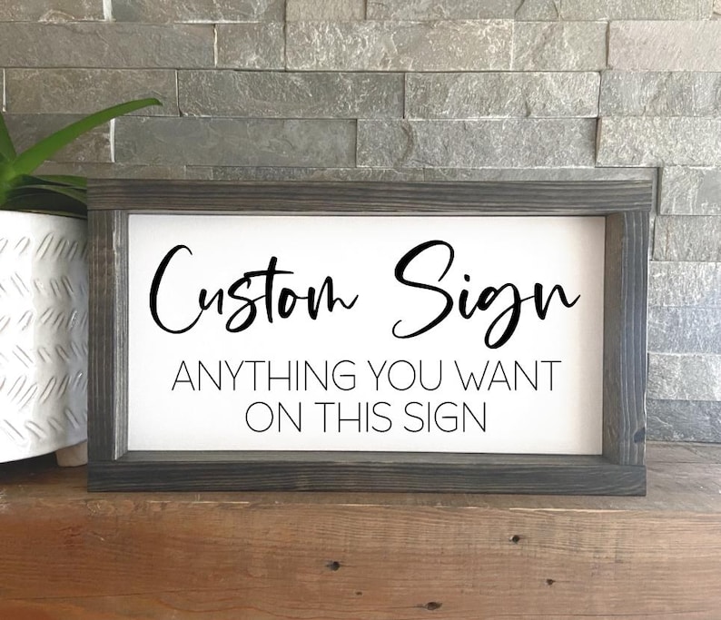 May include: A white wooden sign with a black frame. The sign says "Custom Sign" in a cursive font and "Anything you want on this sign" in a sans-serif font.