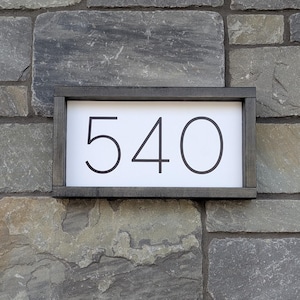 Peut inclure: Panneau d'adresse rectangulaire avec le numéro 540 en noir sur fond blanc. Le panneau est encadré de bois gris foncé et monté sur un mur de pierre.