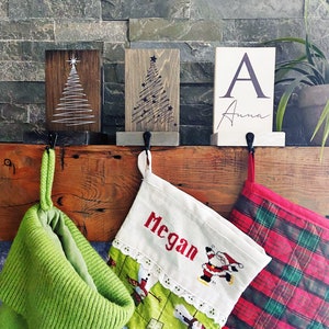 May include: Festive Christmas display with wooden signs depicting Christmas trees and a sign with the letter "A" and the name "Anna". Green, red, and plaid Christmas stockings hang below, creating a holiday atmosphere.