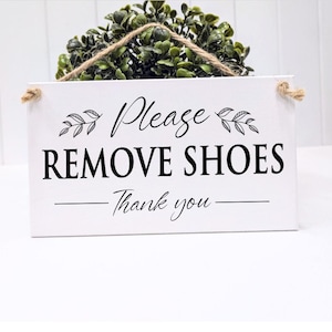 May include: A white rectangular sign with the text "Please REMOVE SHOES - Thank you" in black script. The sign has a decorative leaf design and is suspended by a natural rope. A green plant sits above the sign.