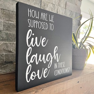 May include: Black wooden sign with white text that reads "How are we supposed to live laugh love in these conditions?"