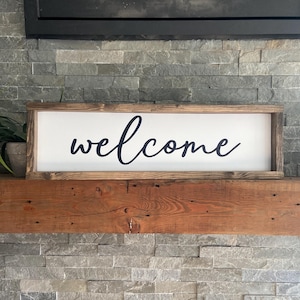 May include: A wooden sign with a white background and black cursive lettering that reads "welcome". The sign is sitting on a wooden mantel above a stone fireplace.