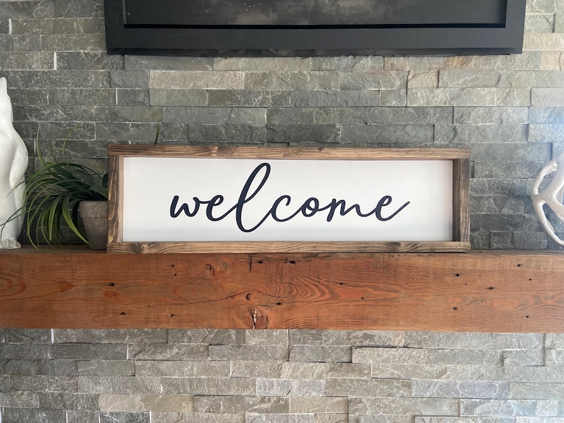 May include: A wooden sign with a white background and black cursive lettering that reads "welcome". The sign is sitting on a wooden mantel above a stone fireplace.