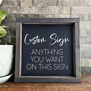 May include: A framed black sign with white text that reads "Custom Sign" and "ANYTHING YOU WANT ON THIS SIGN." The frame is a dark grey colour. A potted plant is visible in the background.
