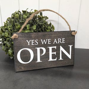 May include: A dark brown wooden sign with the white text "YES WE ARE OPEN". The sign has a rope handle and is set against a backdrop of green foliage.