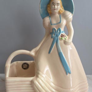 May include: A vintage ceramic figurine of a woman in a long dress and a hat, holding a small bouquet. The figurine is primarily cream-colored with light blue accents on the hat, ribbons, and dress. A basket is attached to the figurine.