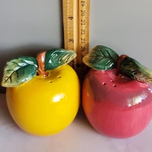 Puede incluir: Un par de saleros y pimenteros de cerámica con forma de manzana. Uno es amarillo, el otro rojo. Ambos tienen detalles de hojas verdes y tallos marrones. Los saleros están sobre una superficie blanca.