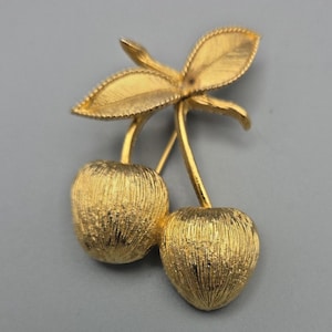 May include: A gold-toned brooch in the shape of two cherries with leaves. The brooch has a textured surface, giving it a vintage look. The cherries are connected by stems and topped with detailed leaves.