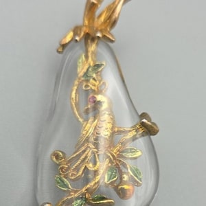 May include: A clear, pear-shaped pendant with a gold-colored bird perched on a branch. The bird has a red gemstone eye. The branch has green leaves and small gold-colored fruit. The top of the pendant has a gold-colored floral design.
