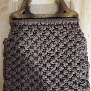 Op de afbeelding: Een bruine macramé handtas met een schildpad handvat. De tas heeft een geweven textuur en een rechthoekige vorm. De handgreep is gebogen en bevestigd aan de bovenkant van de tas. De tas is gemaakt van een zacht, duurzaam materiaal.