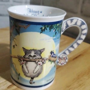 May include: A ceramic mug with a whimsical cat design. The mug features a gray and white cat hanging from a branch, with a full moon and blue sky background. The handle has a blue and white pattern and a small mouse figurine.