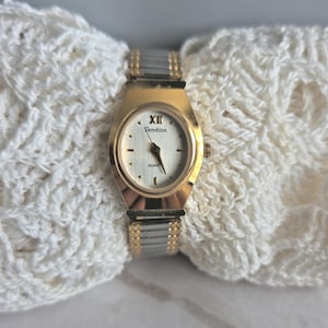 May include: A gold and silver Armitron quartz watch with a white face and black hands. The watch has a gold bezel and a silver and gold band. The watch is displayed on a white, textured surface.