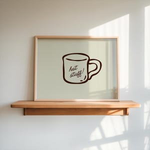 May include: A rectangular framed wall art piece featuring a light beige background and a dark brown line drawing of a coffee mug. The mug illustration includes the handwritten text "hot stuff!" centered within the outline of the cup.