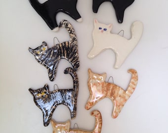May include: A collection of eight ceramic cat ornaments in various colors and patterns, including black, white, brown, and gray. The cats are all in different poses, some sitting, some standing, and some lying down.