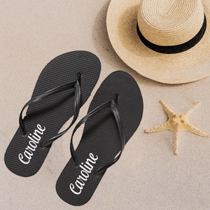 May include: A pair of black flip flops with the name "Caroline" written on each strap. The flip flops are on a sandy surface with a straw hat and a starfish.