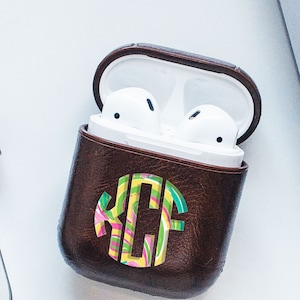May include: A brown leather AirPod case with a colorful monogram design. The case is open, revealing white AirPods inside. The monogram features the letters KCF in a swirl of pink, yellow, green, and blue.