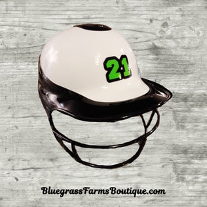May include: White baseball helmet with a black visor and a black cage. The helmet has a green number 21 on the front.