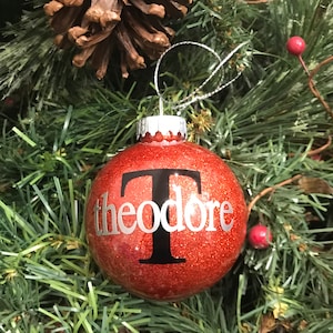 May include: A red glitter Christmas ornament with the name "Theodore" in silver script and a black "T". The ornament hangs from a silver string and is surrounded by green pine needles and red berries.
