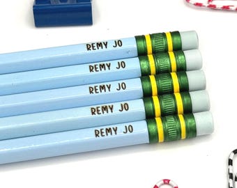 May include: Five light blue pencils arranged horizontally, each featuring the name REMY JO printed in black. The pencils have metallic green and yellow ferrules with white erasers. A blue binder clip and several patterned paper clips are visible.