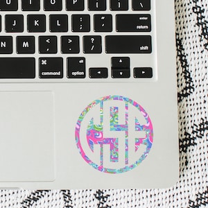 May include: A colorful monogram sticker on a laptop. The sticker features a circular design with a white outline and a pattern of pink, blue, and green swirls. The laptop keyboard is visible.