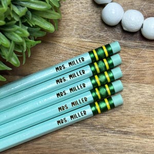 May include: Five light blue pencils with the text "MRS. MILLER" printed on them. The pencils have green and yellow accents near the eraser end. The pencils are arranged diagonally on a wooden surface.
