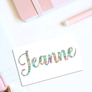 May include: A white card with the name "Jeanne" written in a colorful, patterned font.