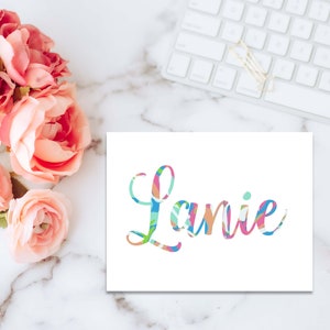 May include: A white card with the name "Lanie" written in colorful, stylized lettering. The letters are filled with a pattern of pink, blue, green, and orange.