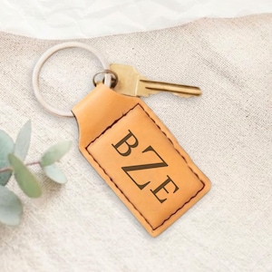 May include: A rectangular tan leather keychain featuring a metal split ring and a brass-colored key. The leather tag is stitched along the perimeter and displays the embossed capital letters BZE in a dark serif font centered on the front surface.