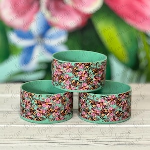 May include: Three teal blue coasters with a floral and butterfly design. The coasters feature a pattern of pink and yellow flowers with orange and brown butterflies.