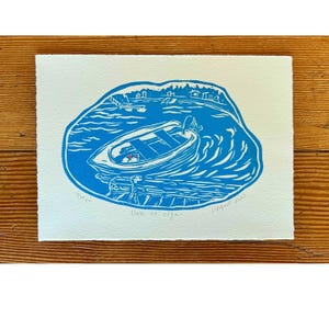 May include: A blue and white print of a boat on the water, with a shoreline in the background. The print is titled "Dock at Olga" and includes the artist's signature and the number 4/20. The boat is docked at a pier.