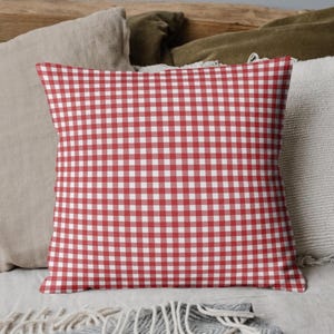 May include: A square decorative pillow with a red and white gingham pattern. The pillow is the focal point, resting among other pillows in neutral tones. The pillow is likely used for home decor.