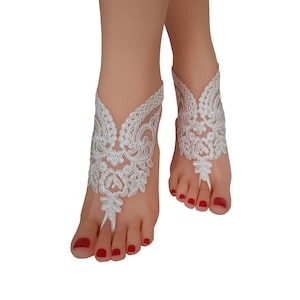 May include: White lace barefoot sandals. The sandals feature an intricate floral design, with a toe loop and ankle strap. The delicate lace is detailed with floral patterns. The sandals are designed to be worn on the feet.