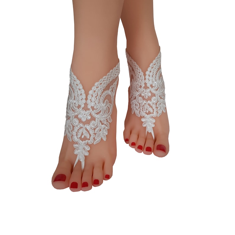 May include: White lace barefoot sandals. The sandals feature an intricate floral design, with a toe loop and ankle strap. The delicate lace is detailed with floral patterns. The sandals are designed to be worn on the feet.