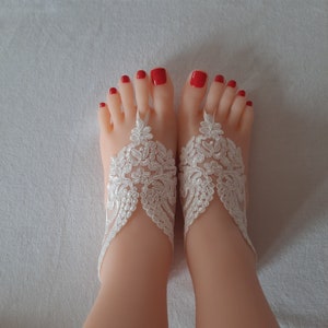 May include: A pair of feet with red nail polish, adorned with white lace barefoot sandals. The sandals feature intricate floral patterns and delicate straps, perfect for a beach wedding or special occasion.