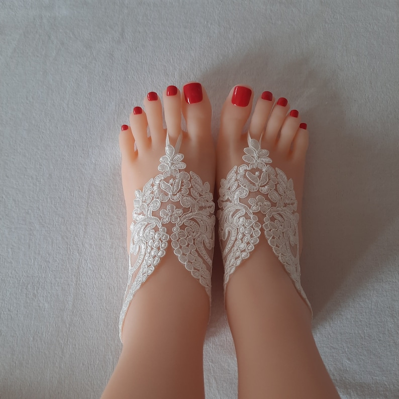 May include: A pair of feet with red nail polish, adorned with white lace barefoot sandals. The sandals feature intricate floral patterns and delicate straps, perfect for a beach wedding or special occasion.