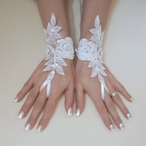 Peut inclure: Une paire de gants sans doigts en dentelle blanche avec des motifs floraux et un détail de ruban satiné. Les gants sont portés sur une paire de mains avec un vernis à ongles blanc.