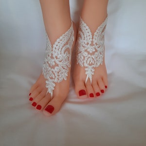 May include: White lace barefoot sandals with floral and leaf patterns. The sandals are designed to be worn on the feet, with a toe loop and ankle strap. The toenails are painted red.