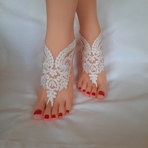 May include: White lace barefoot sandals with floral and scrollwork designs. The sandals feature a toe loop and ankle cuff, perfect for a beach wedding or special occasion. The feet have red nail polish.