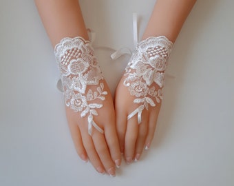Puede incluir: Un par de guantes de encaje blanco sin dedos con un intrincado patrón de bordado floral. Los guantes se aseguran alrededor de los dedos medios con finas lazadas de cinta blanca y presentan bordes festoneados en las muñecas con cintas a juego.