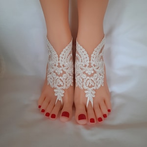 May include: White lace barefoot sandals with floral and leaf patterns. The sandals feature a toe loop and ankle cuff design, perfect for a beach wedding or special occasion. The sandals are displayed on a pair of feet with red nail polish.